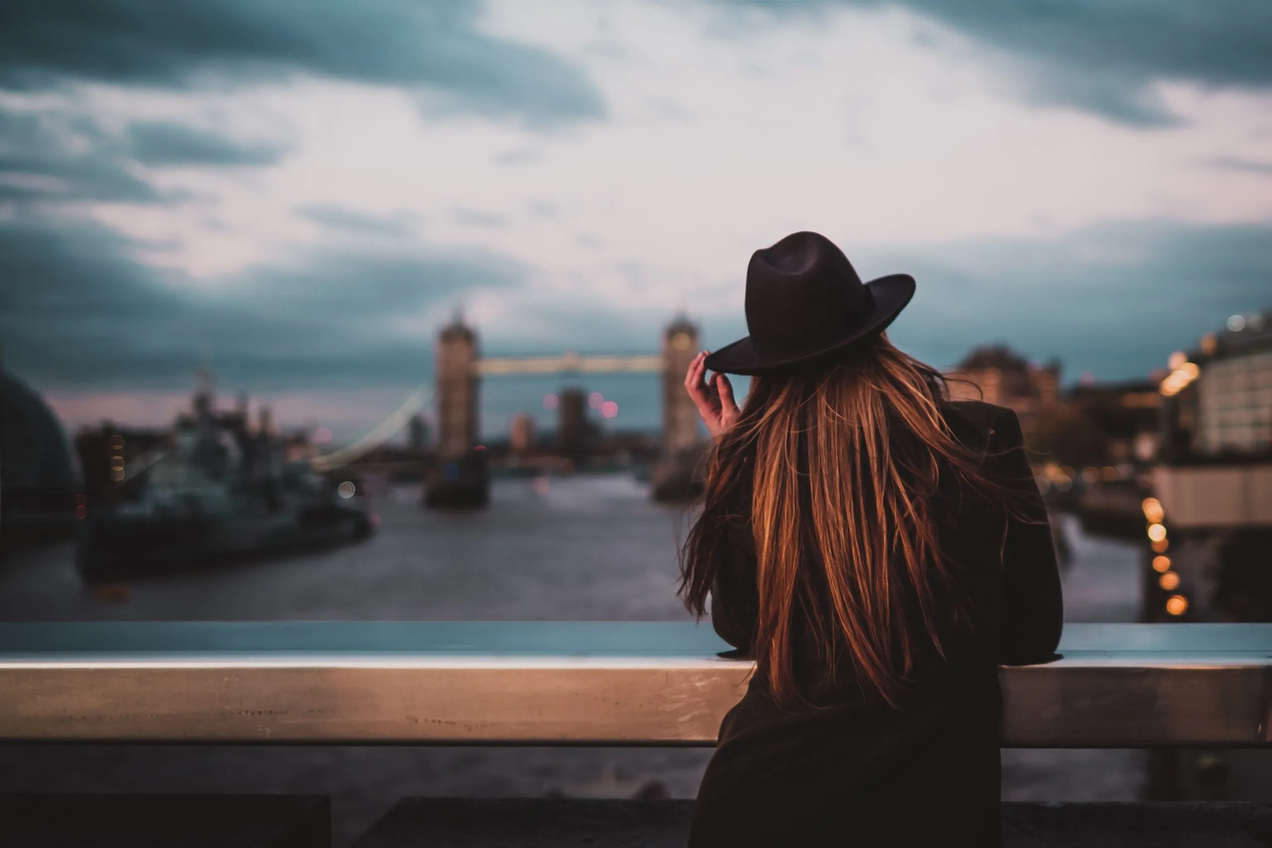 A woman in a black hat gazes at Tower Bridge in London during twilight.