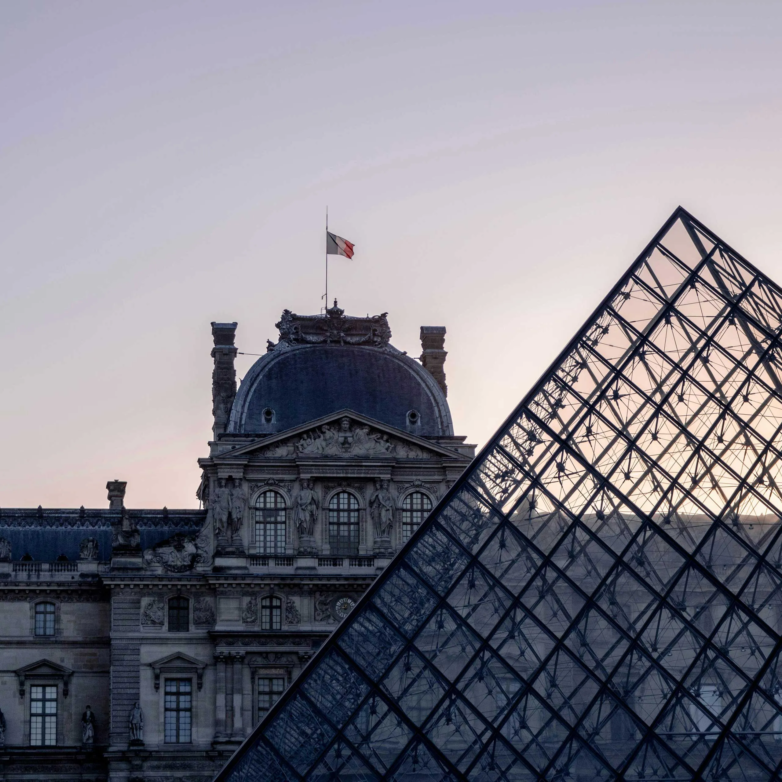 The Louvre's historic facade and modern glass pyramid are juxtaposed at dusk, with a French flag atop.
