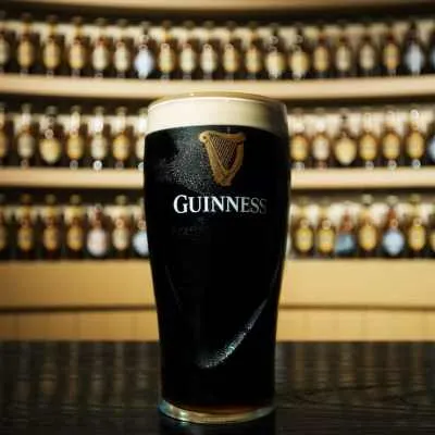 A pint of Guinness stout with a creamy head, set against a backdrop of bottles on shelves.