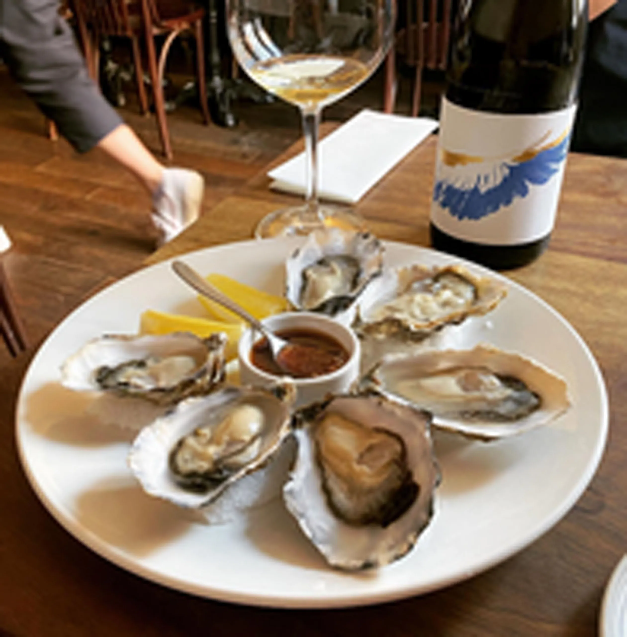 Plate of fresh oysters with sauce, lemon wedges, white wine, and bottle on a wooden restaurant table.
