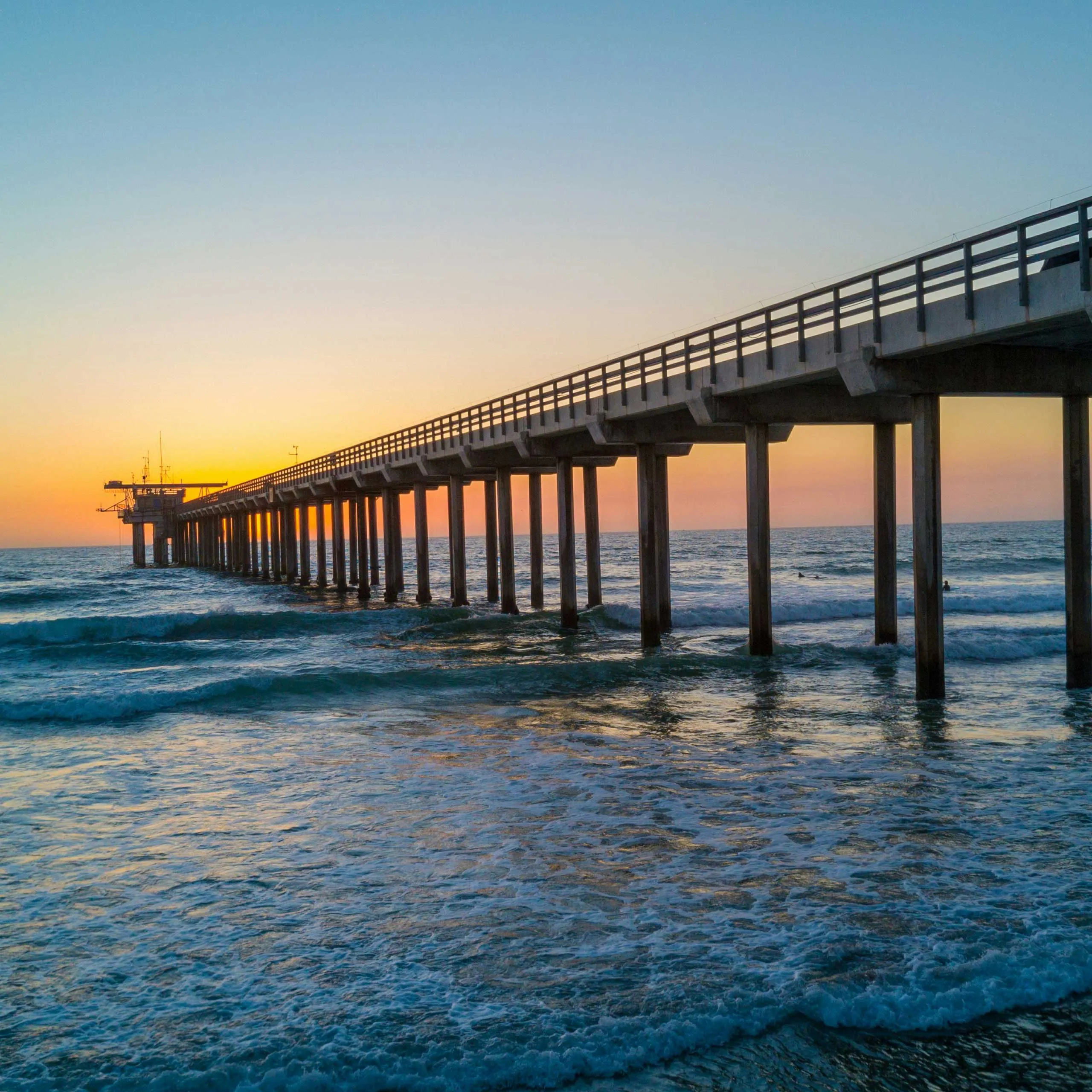 A long pier extends over ocean waves at sunset, with warm hues blending into the horizon.