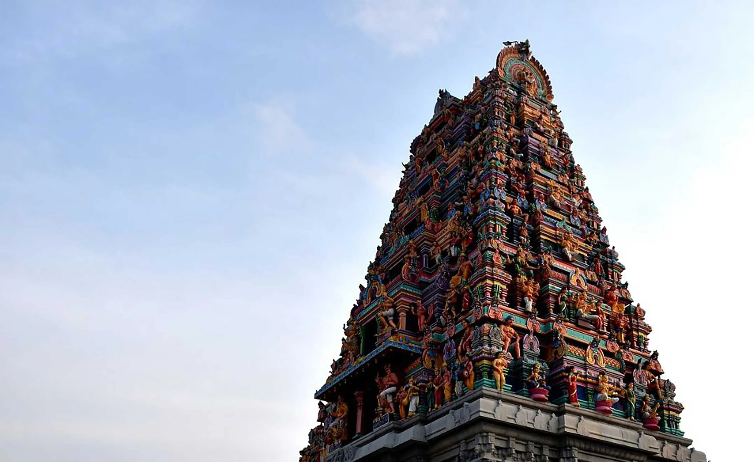 A vibrant Hindu temple tower adorned with intricate sculptures rises against a clear blue sky.