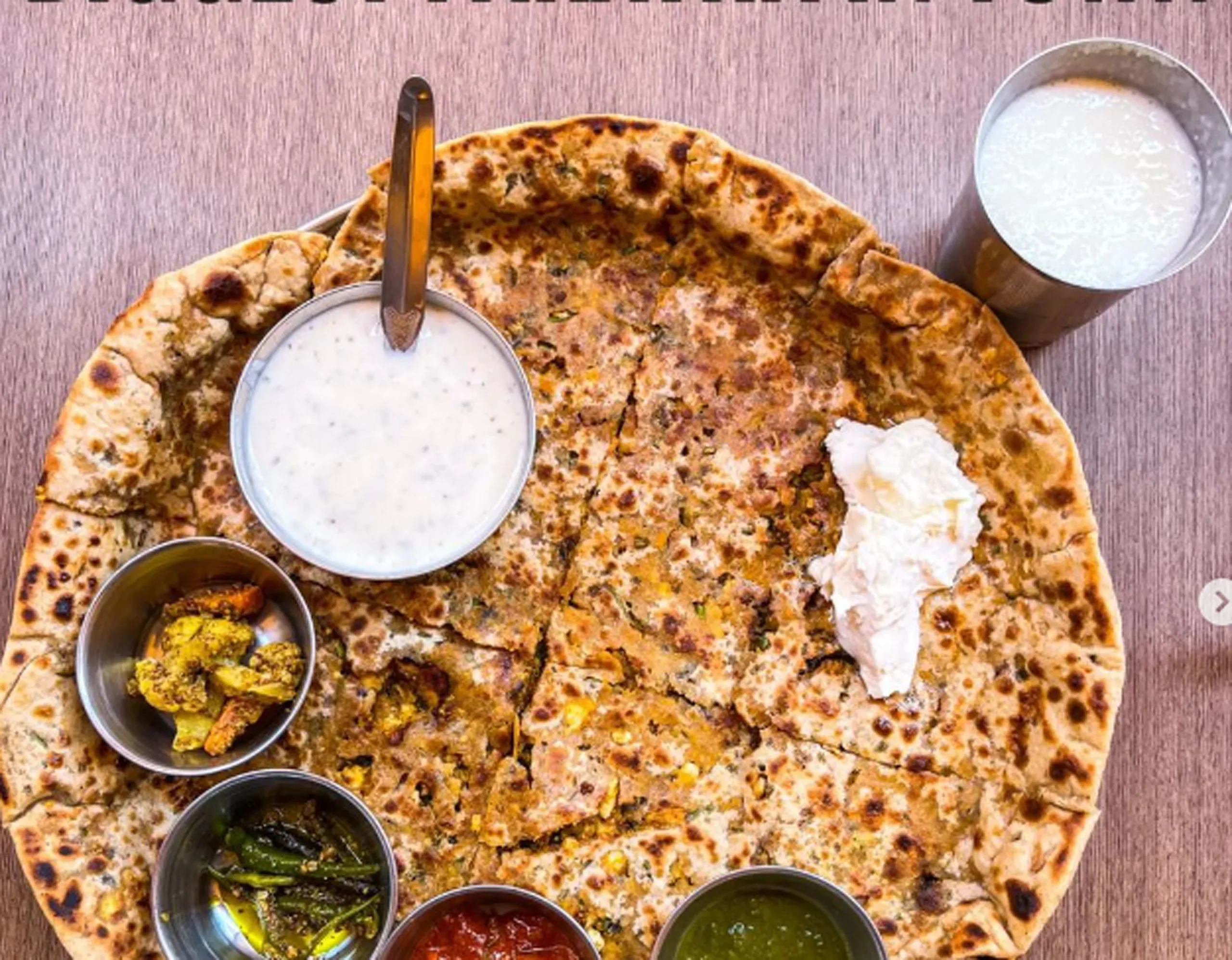 A large stuffed paratha served with curd, butter, pickles, chutneys, and a glass of buttermilk.