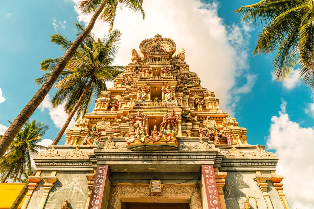A vibrant Hindu temple tower adorned with intricate carvings, framed by palm trees and a blue sky.