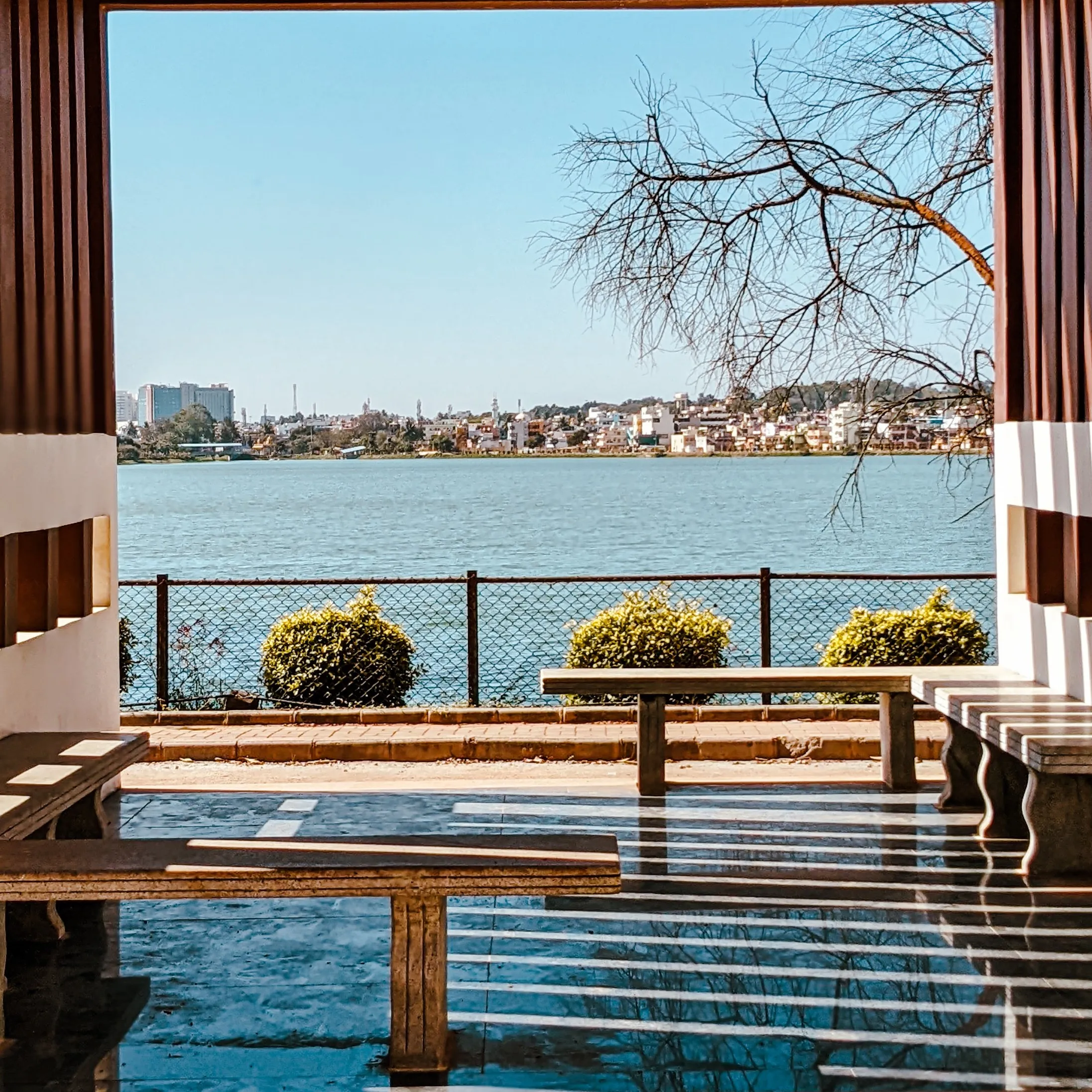 A serene lakeside view framed by wooden panels, benches, and a tree, with a cityscape in the distance.