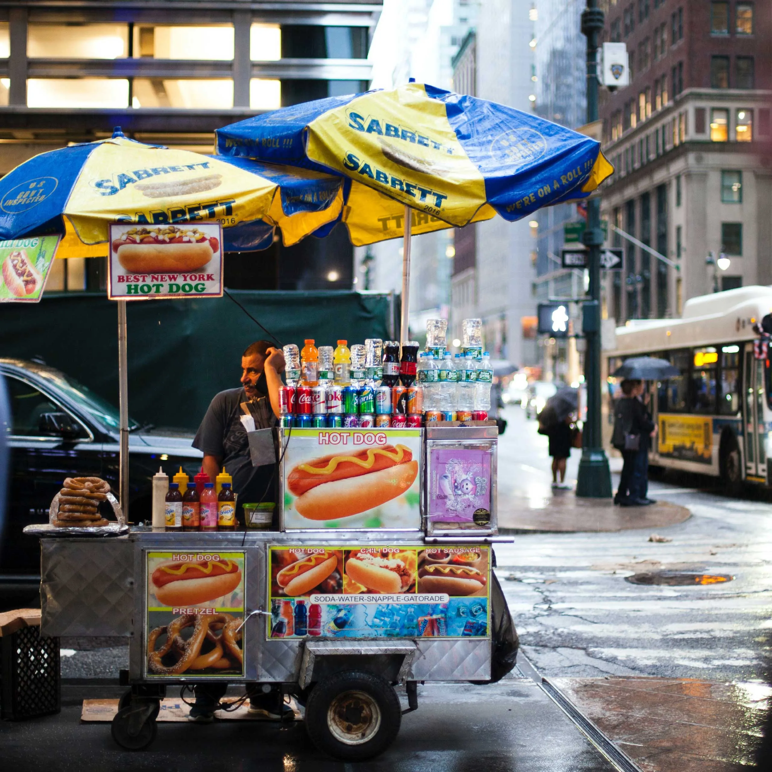A New York City hot dog cart with Sabrett umbrellas, beverages, and condiments on a rainy street.