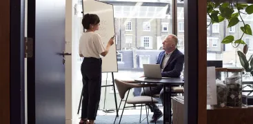 A woman presents at a whiteboard to a seated man in a modern office with large windows.