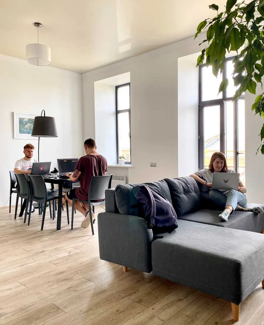 A modern living space with people working at a table and lounging on a gray sectional sofa.