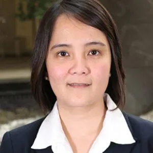 A woman wearing a dark blazer and white collared shirt stands outdoors near a blurred background.