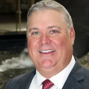 A man in a suit and red tie stands outdoors near a fountain, with a professional appearance.