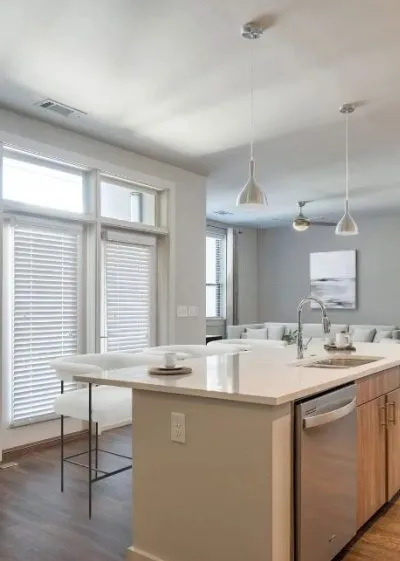 Modern kitchen with pendant lights, white island, stainless steel dishwasher, and large windows.