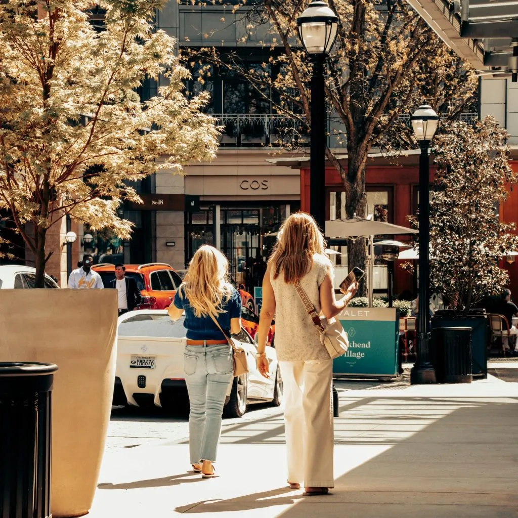 Two women walk in a sunny urban shopping area, surrounded by trees, shops, and parked cars.