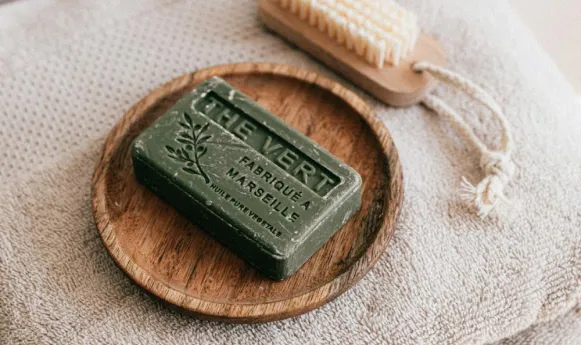 A green soap bar labeled "Thé Vert" rests on a wooden dish beside a brush and towel.