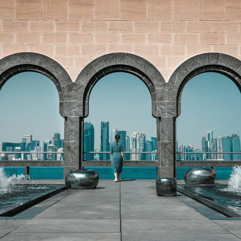 A woman stands framed by arches, overlooking a modern city skyline across the water.