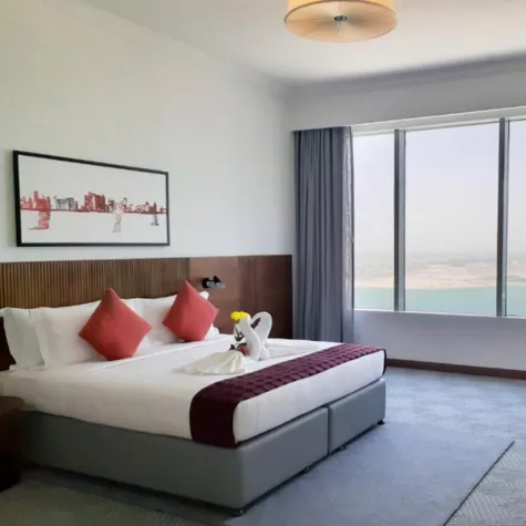 Modern hotel room with a large bed, red accents, wall art, and a window overlooking the sea.
