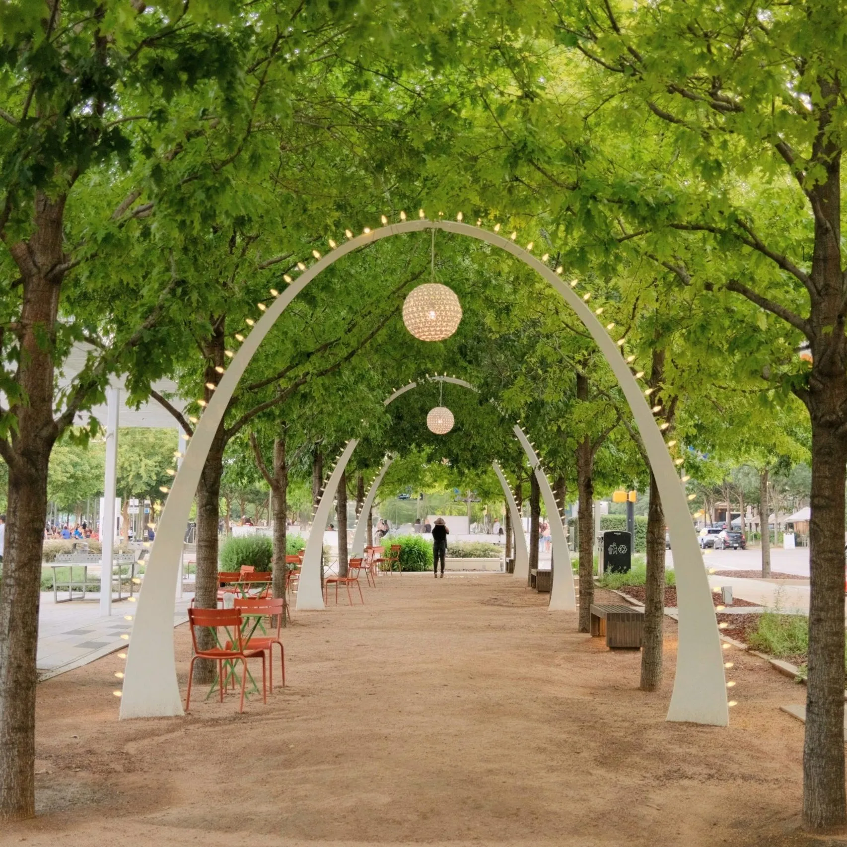 A tree-lined walkway features arched lights, hanging globes, and scattered seating on a dirt path.