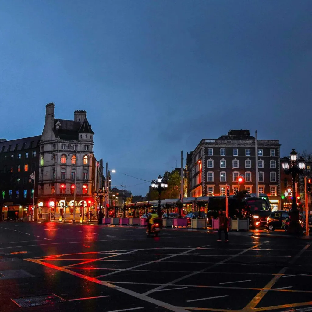 A rainy evening cityscape with illuminated buildings, streetlights, and light reflections on wet roads.
