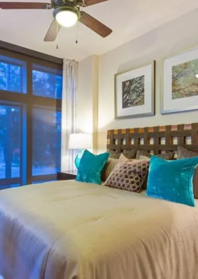 A modern bedroom with teal accent pillows, wooden headboard, and large windows with natural light.