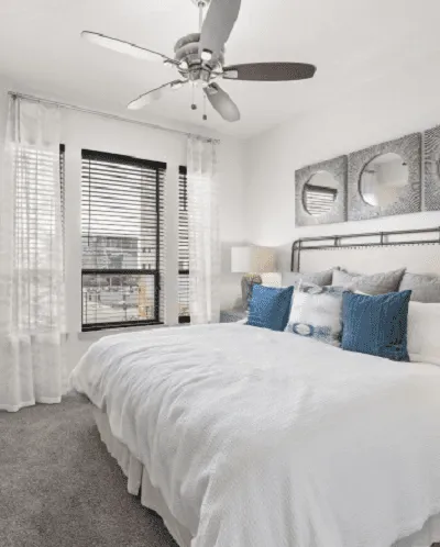 Bright bedroom with modern decor, white bedding, blue accents, and a ceiling fan.