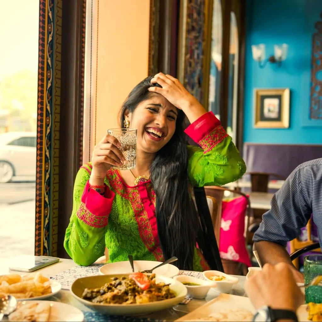A woman in vibrant attire enjoys a meal at a colorful restaurant, holding a glass in her hand.
