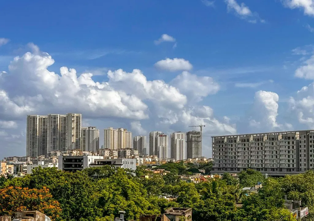 Urban skyline with high-rise buildings, lush greenery, and scattered clouds under a bright blue sky.