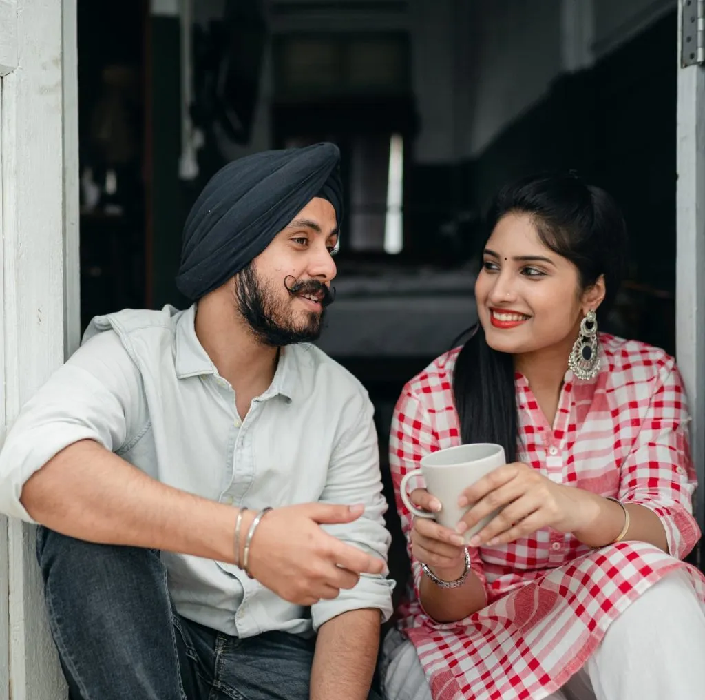 A man in a turban and a woman in a checkered outfit sit together, conversing while holding a cup.