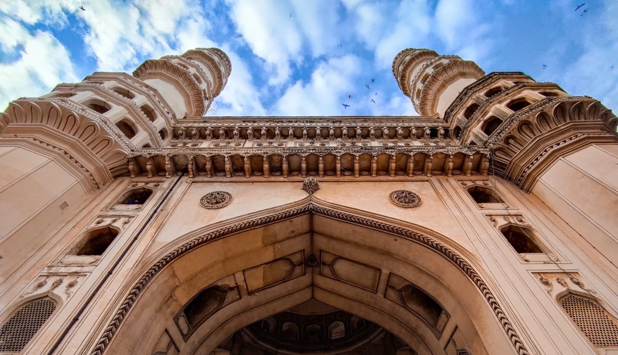 Low-angle view of Charminar's intricate architecture against a vibrant blue sky with scattered clouds.