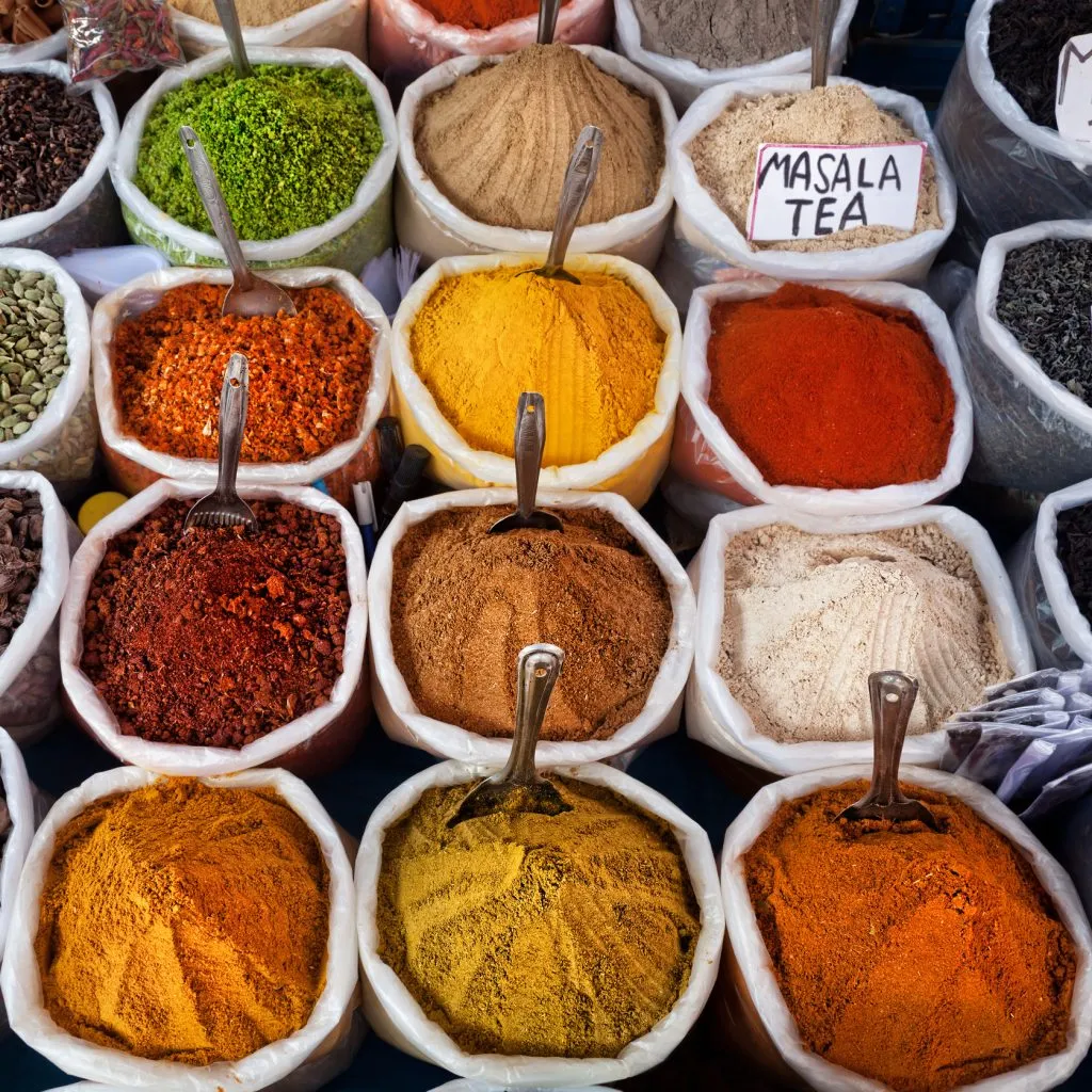 Colorful spices displayed in sacks with spoons at a market, including a labeled "Masala Tea" mix.