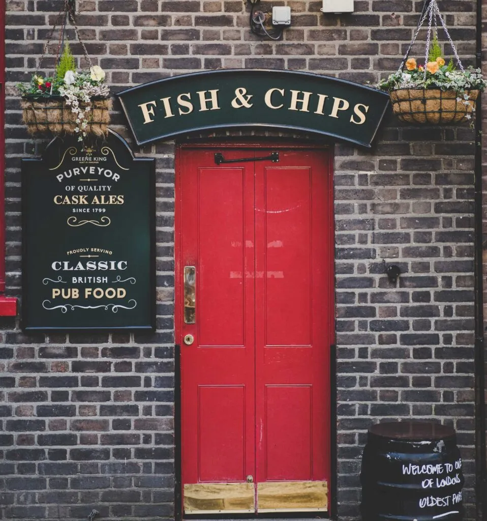 A red door with signage for a British pub serving fish, chips, and cask ales, flanked by flowers.