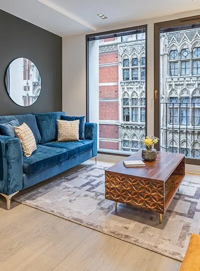 Modern living room with a velvet blue sofa, geometric wood table, and large windows.