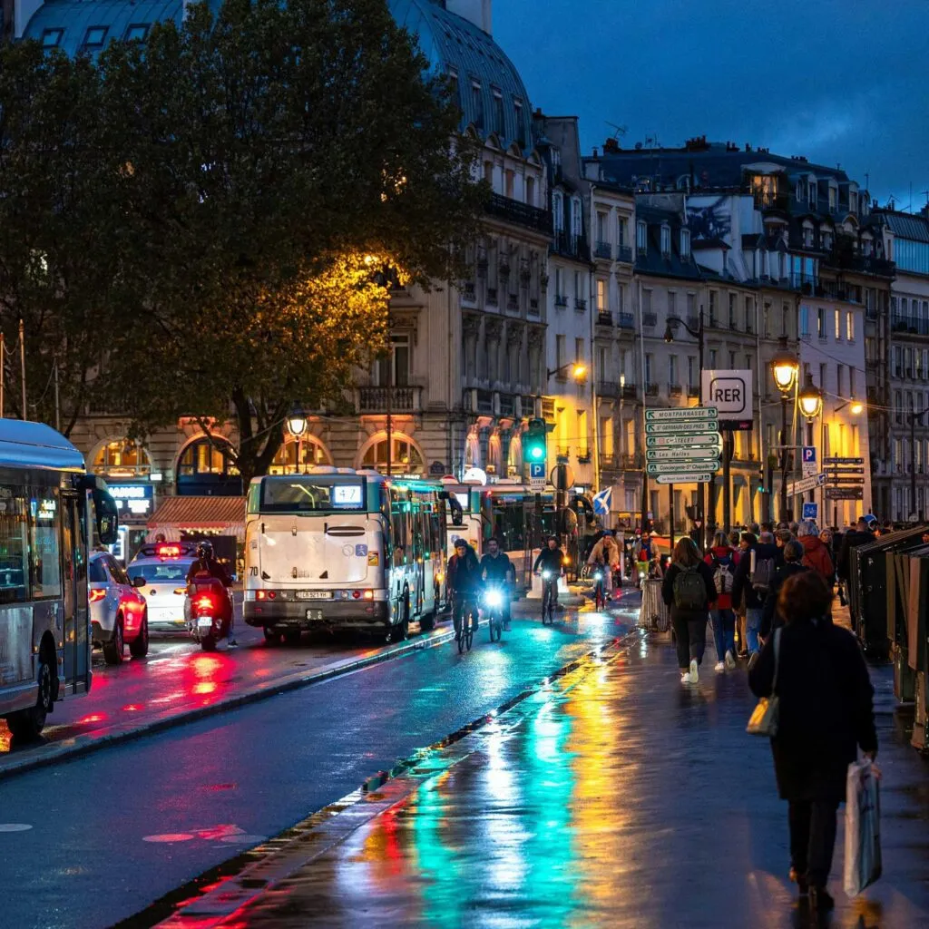 A bustling city street at night with buses, cyclists, pedestrians, and colorful reflections on wet pavement.