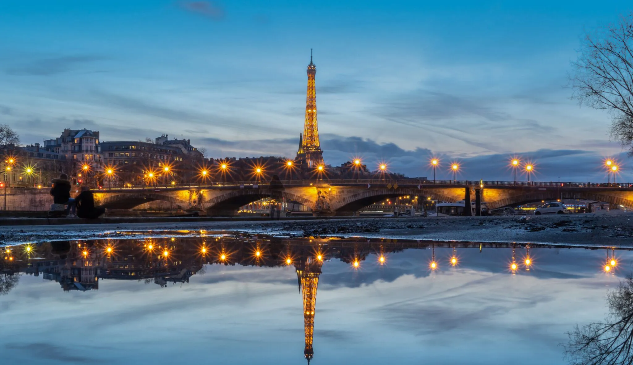 The Eiffel Tower and bridge lights reflect in a calm water surface at dusk, creating a serene scene.