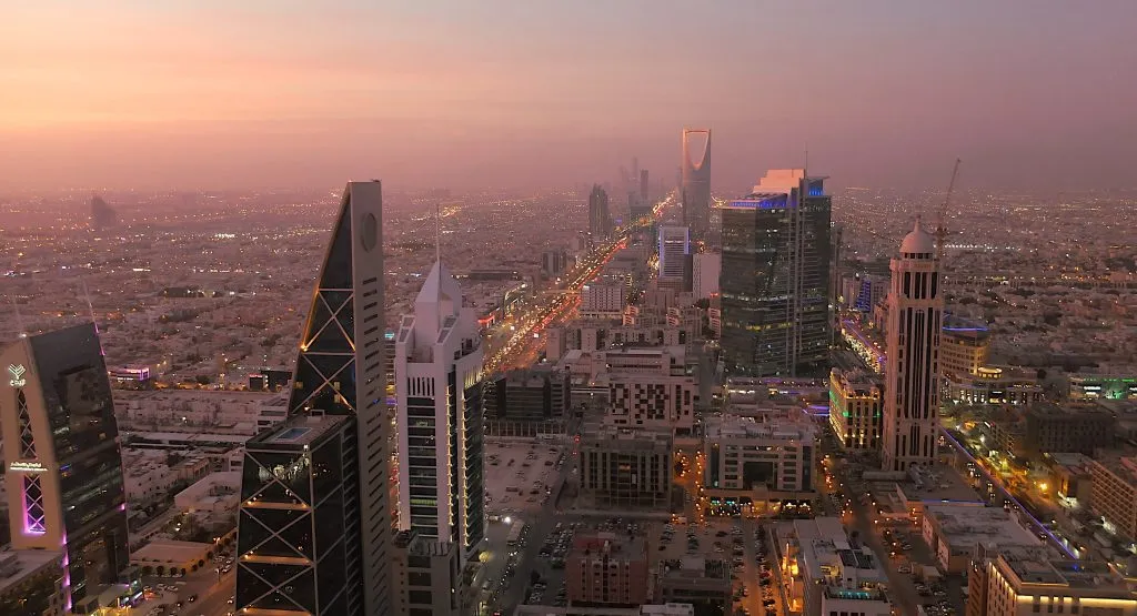 A vibrant cityscape of Riyadh at sunset, showcasing modern skyscrapers and illuminated streets.