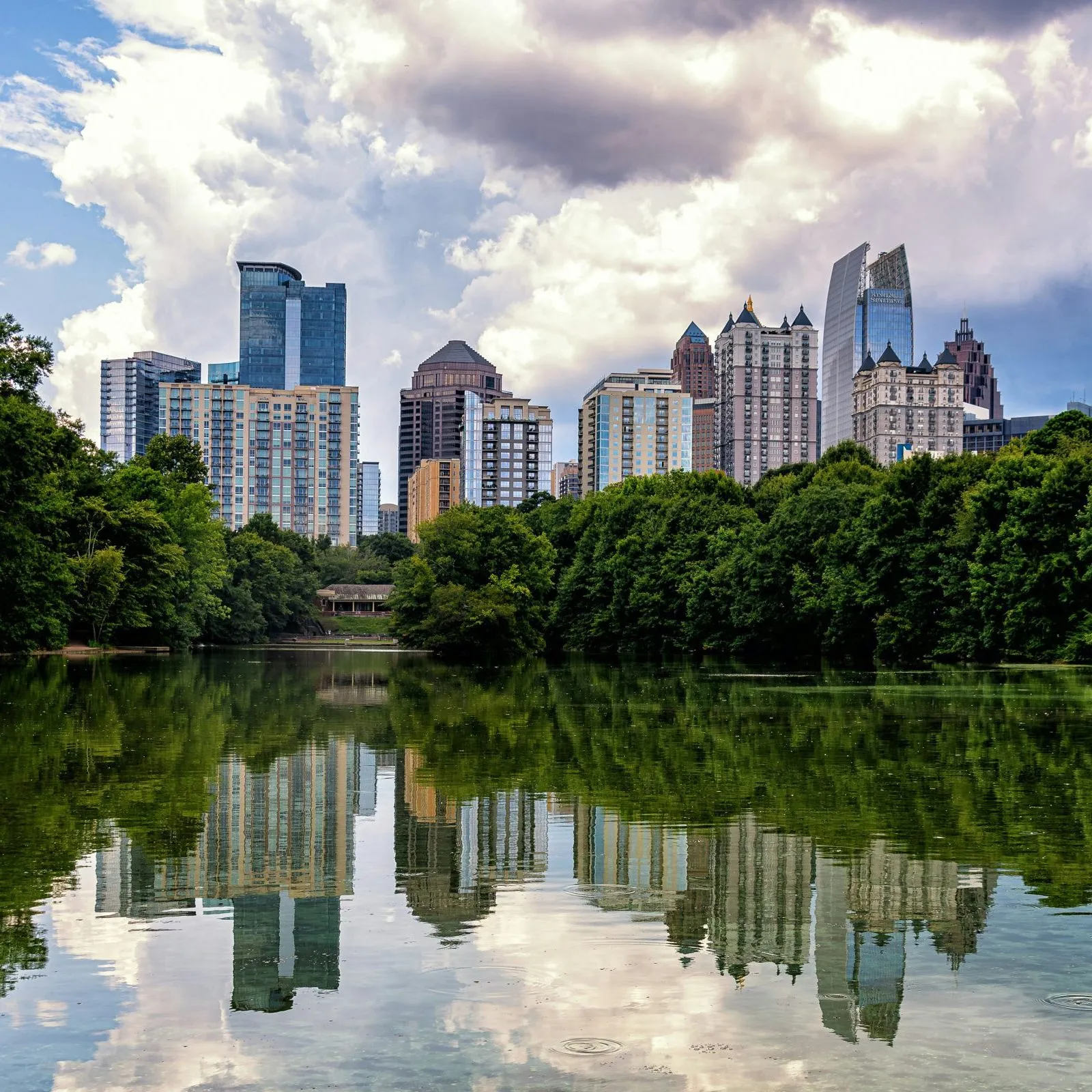 A city skyline reflects on a calm lake, framed by lush green trees under a cloudy sky.