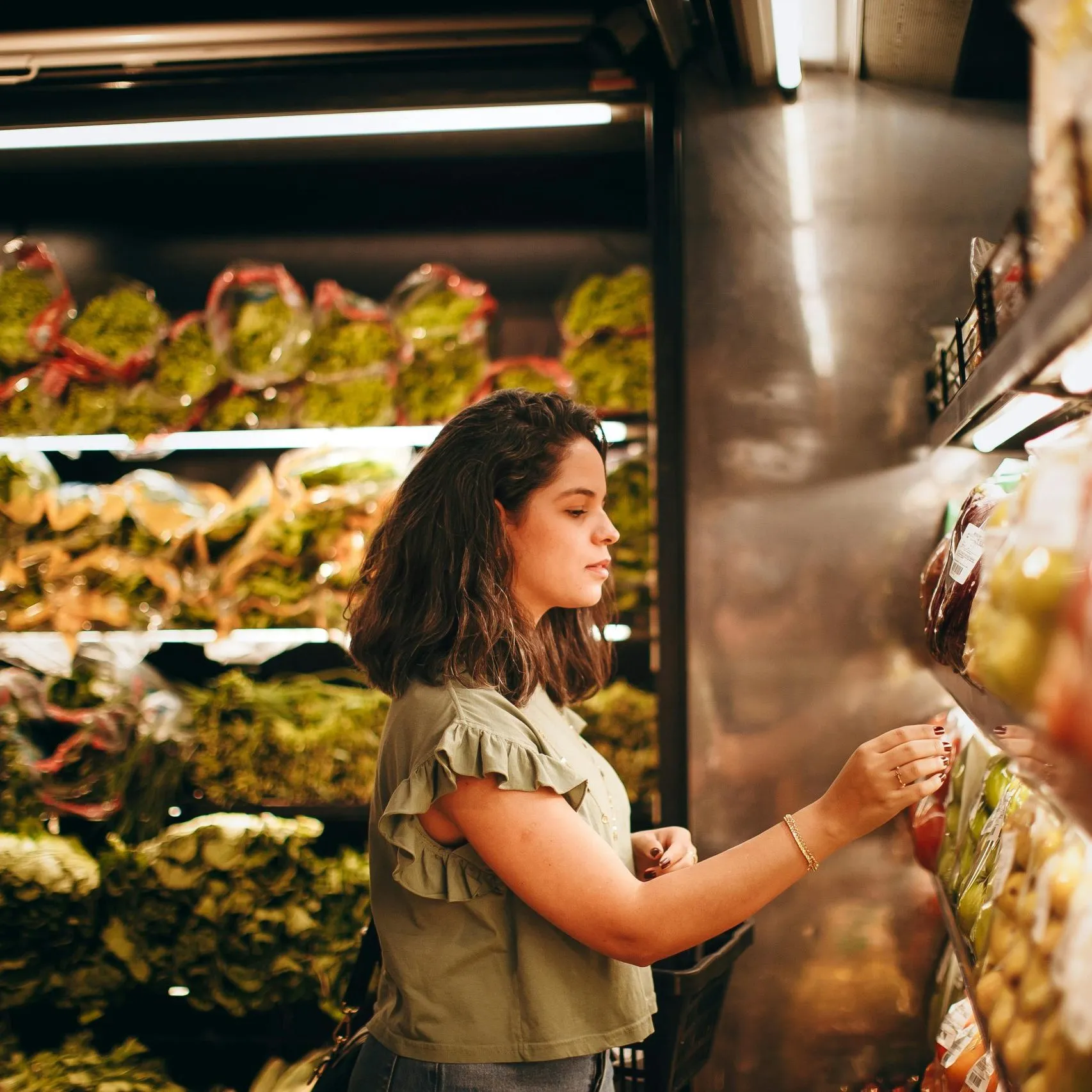 A woman wearing a green ruffled top shops for produce in a well-lit grocery store aisle.