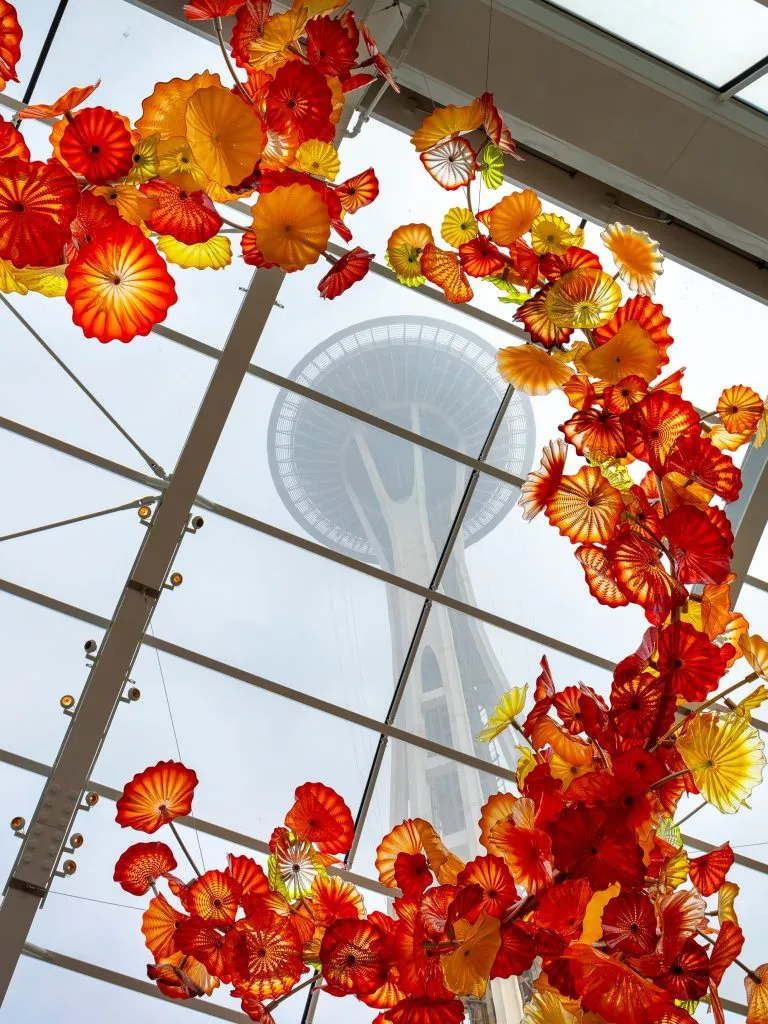 Vibrant glass art installation frames the Space Needle through a glass ceiling on a cloudy day.