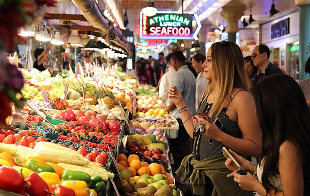 A vibrant market scene with fresh produce and seafood, bustling with shoppers under neon signage.