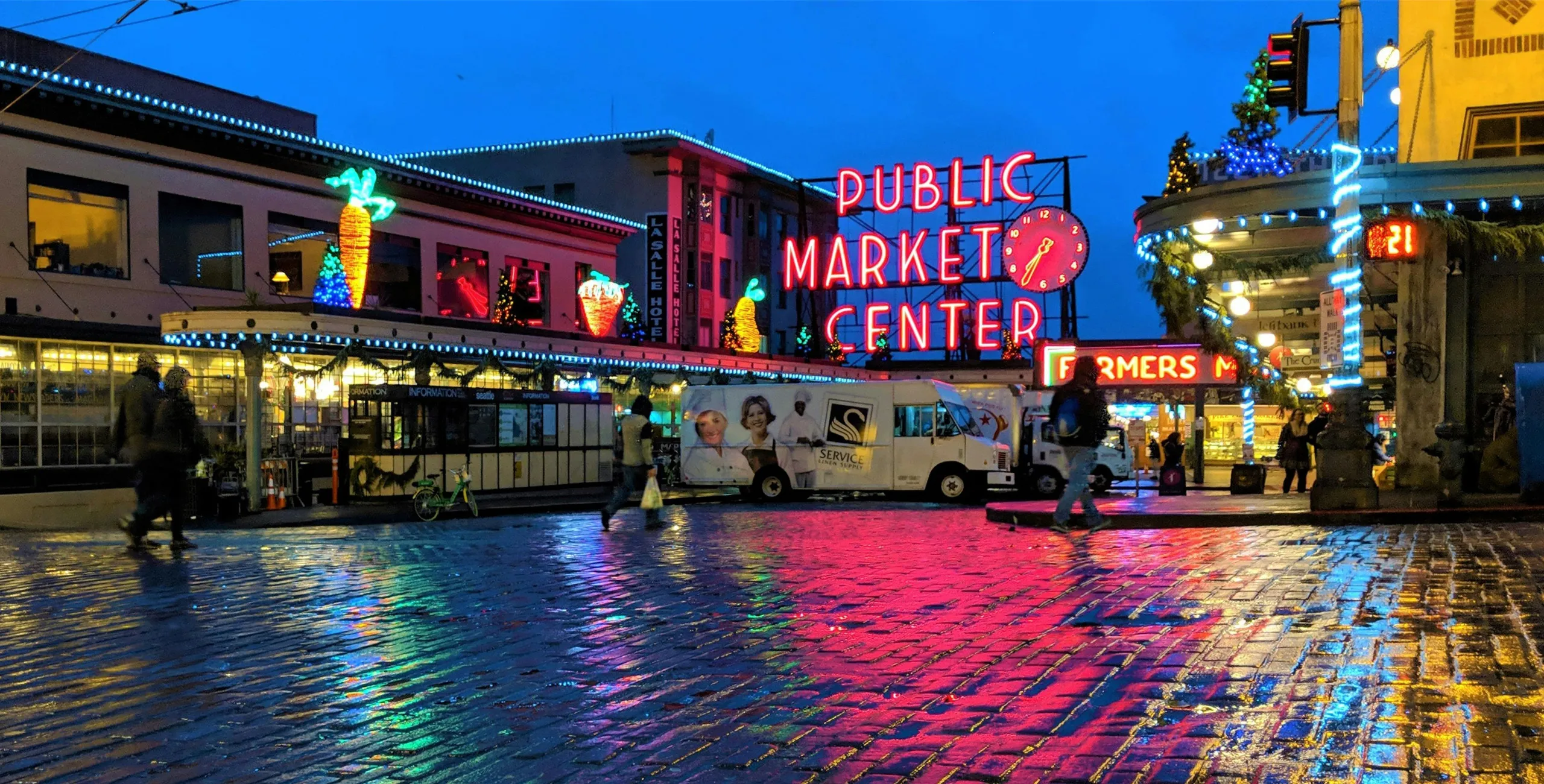 Pike Place Market illuminated at night, reflecting vibrant neon lights on wet cobblestone streets.