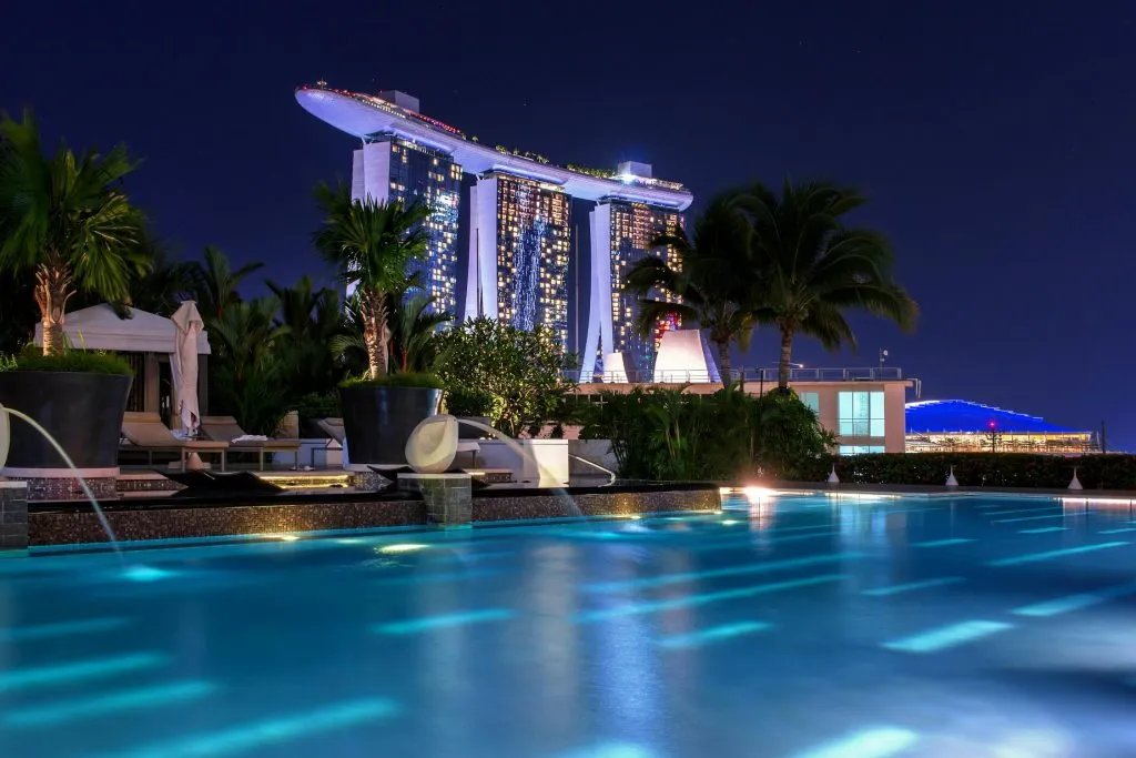 Marina Bay Sands illuminated at night, viewed from a luxurious pool surrounded by tropical greenery.