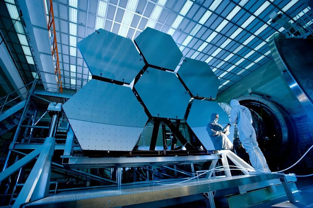 Engineers in cleanroom suits inspect hexagonal mirror segments in a high-tech laboratory setting.
