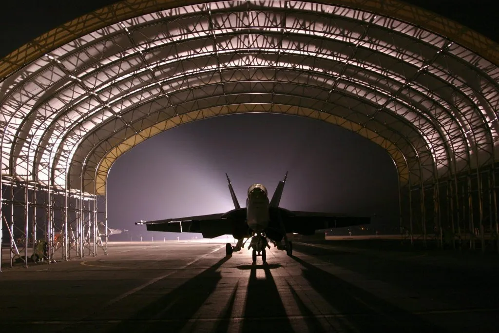 A fighter jet is silhouetted under a large, illuminated hangar at night, emphasizing its sleek form.