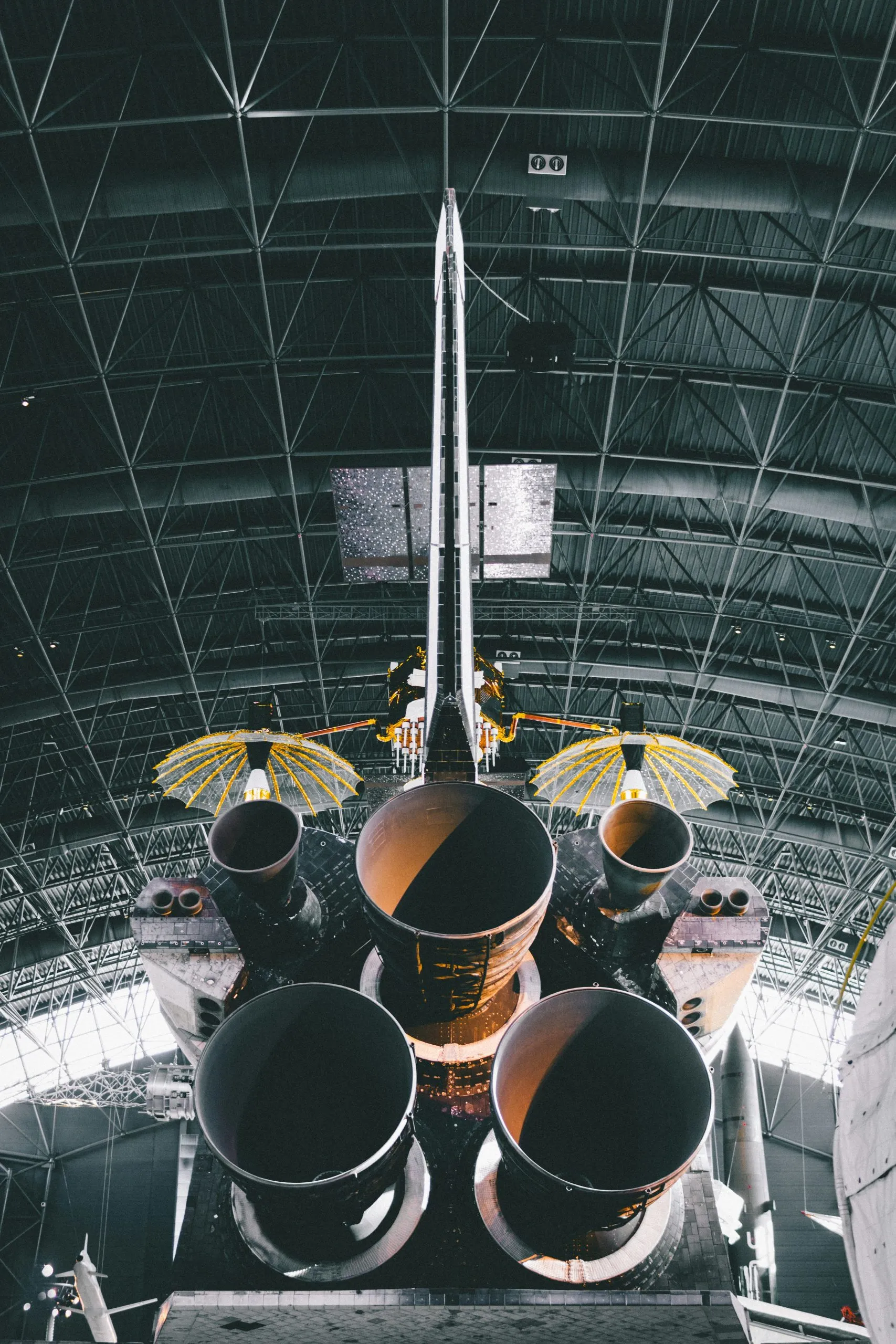 Space shuttle rear view with rocket engines, solar panels, and wing-like structures in a hangar.