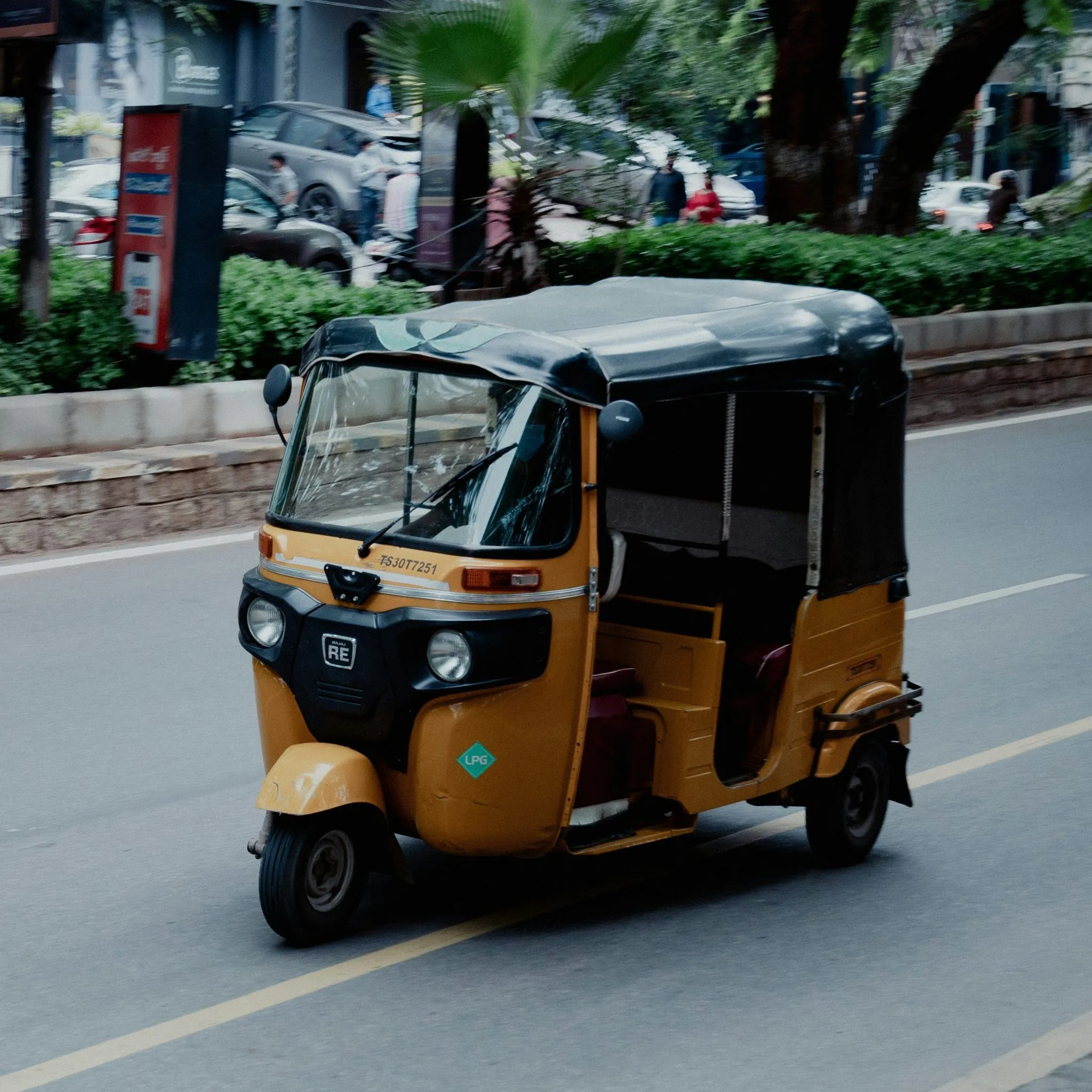 A yellow auto-rickshaw drives on a city street, surrounded by greenery and urban structures.