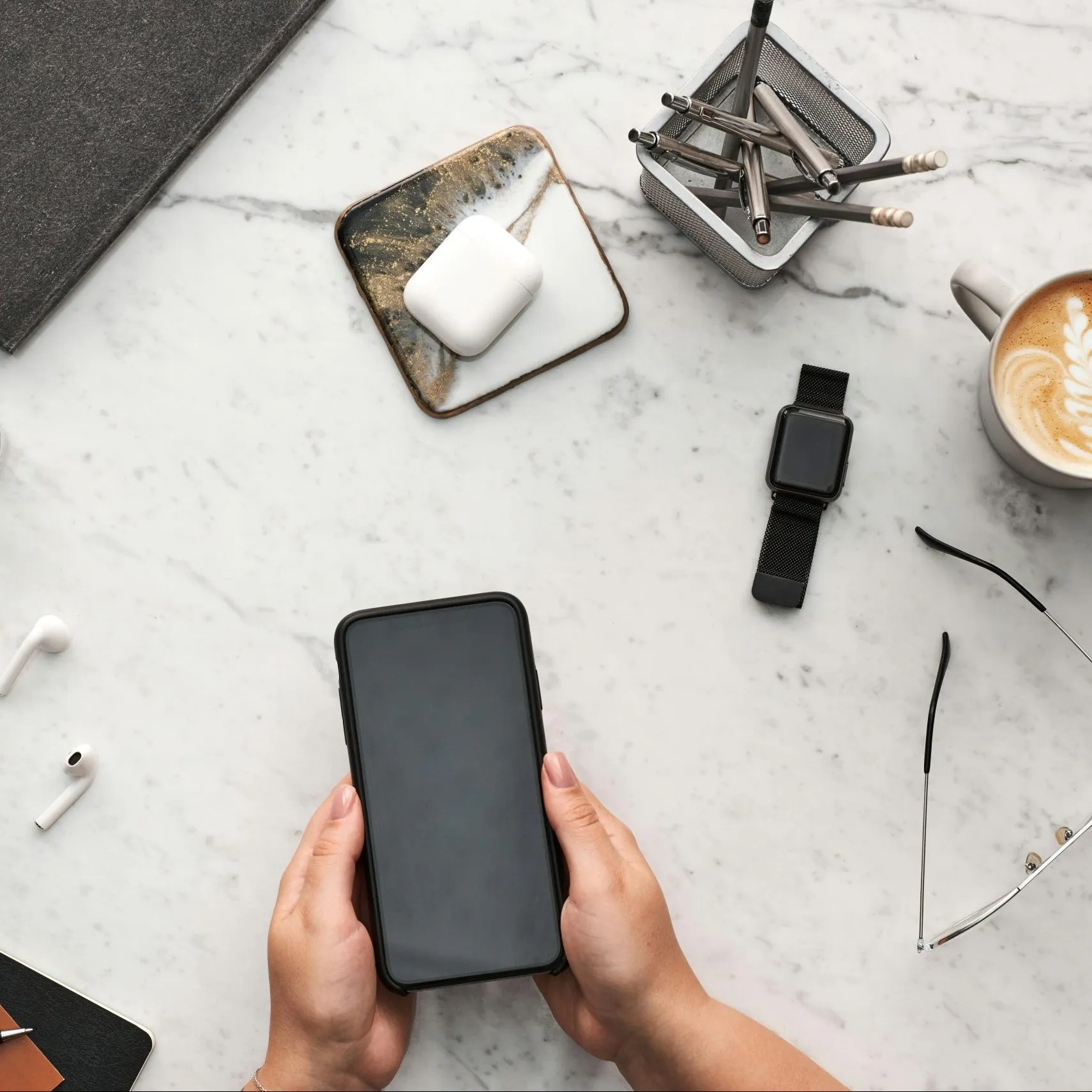 Hands holding a smartphone on a marble desk with AirPods, smartwatch, pens, and coffee nearby.