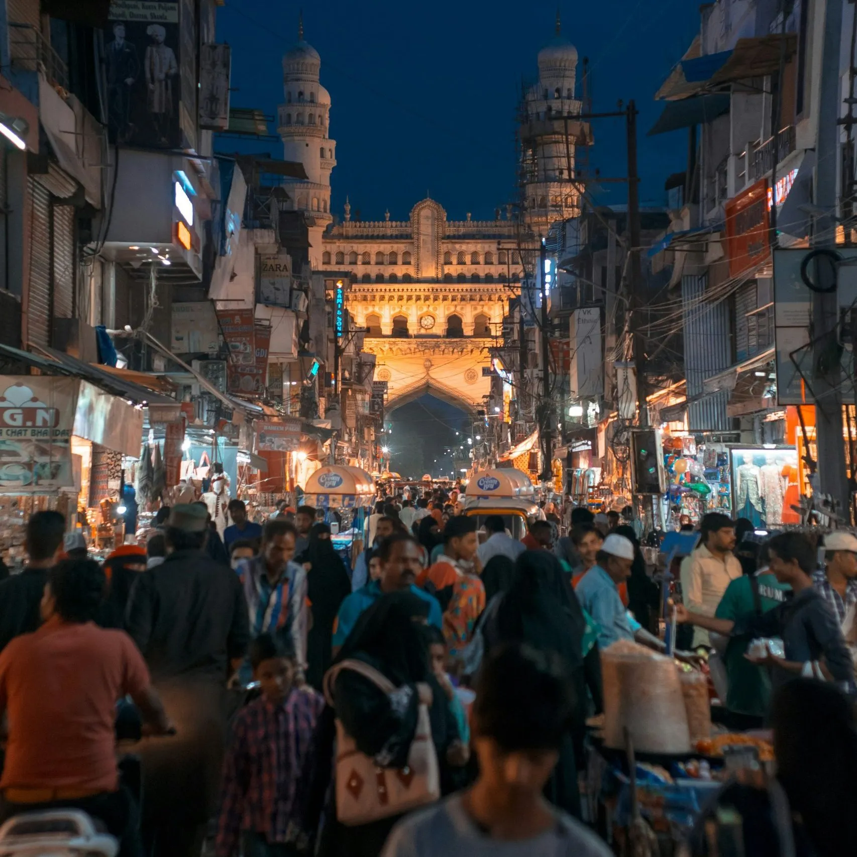 A bustling market street leads to the illuminated Charminar, surrounded by vibrant shops and activity.