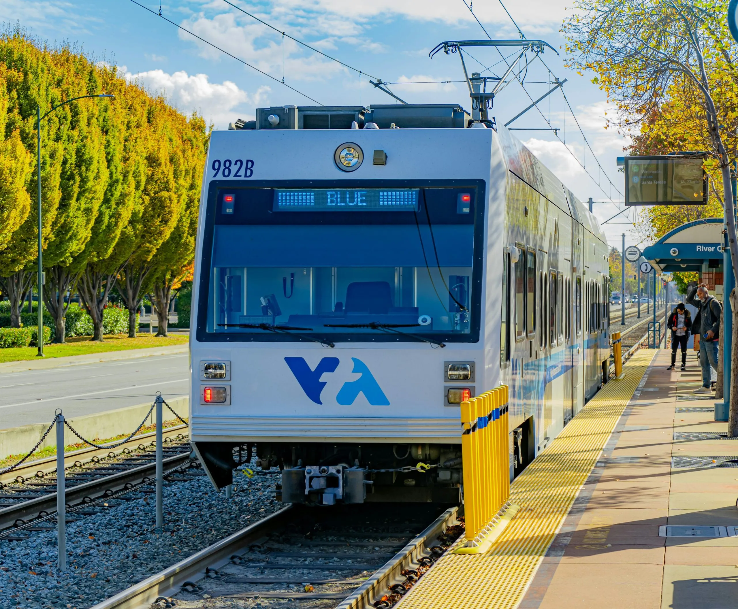 A VTA light rail train labeled "BLUE" is stopped at a station platform surrounded by autumn trees.