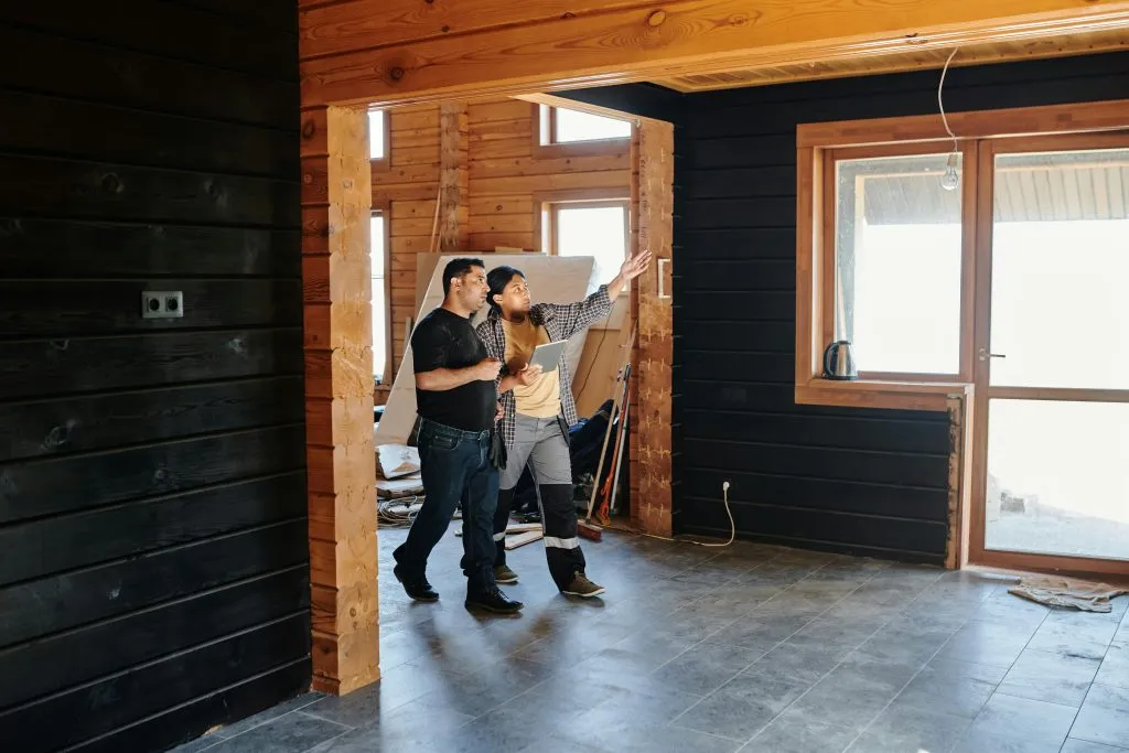 Two individuals discuss interior renovations in a partially completed wooden house with large windows.