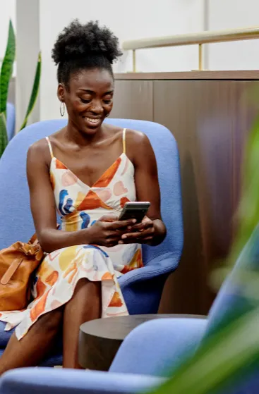 A woman in a colorful dress sits on a blue chair, holding a phone with a bag beside her.