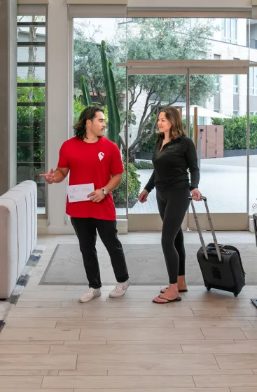 A person in a red shirt assists a traveler with luggage in a modern building lobby.