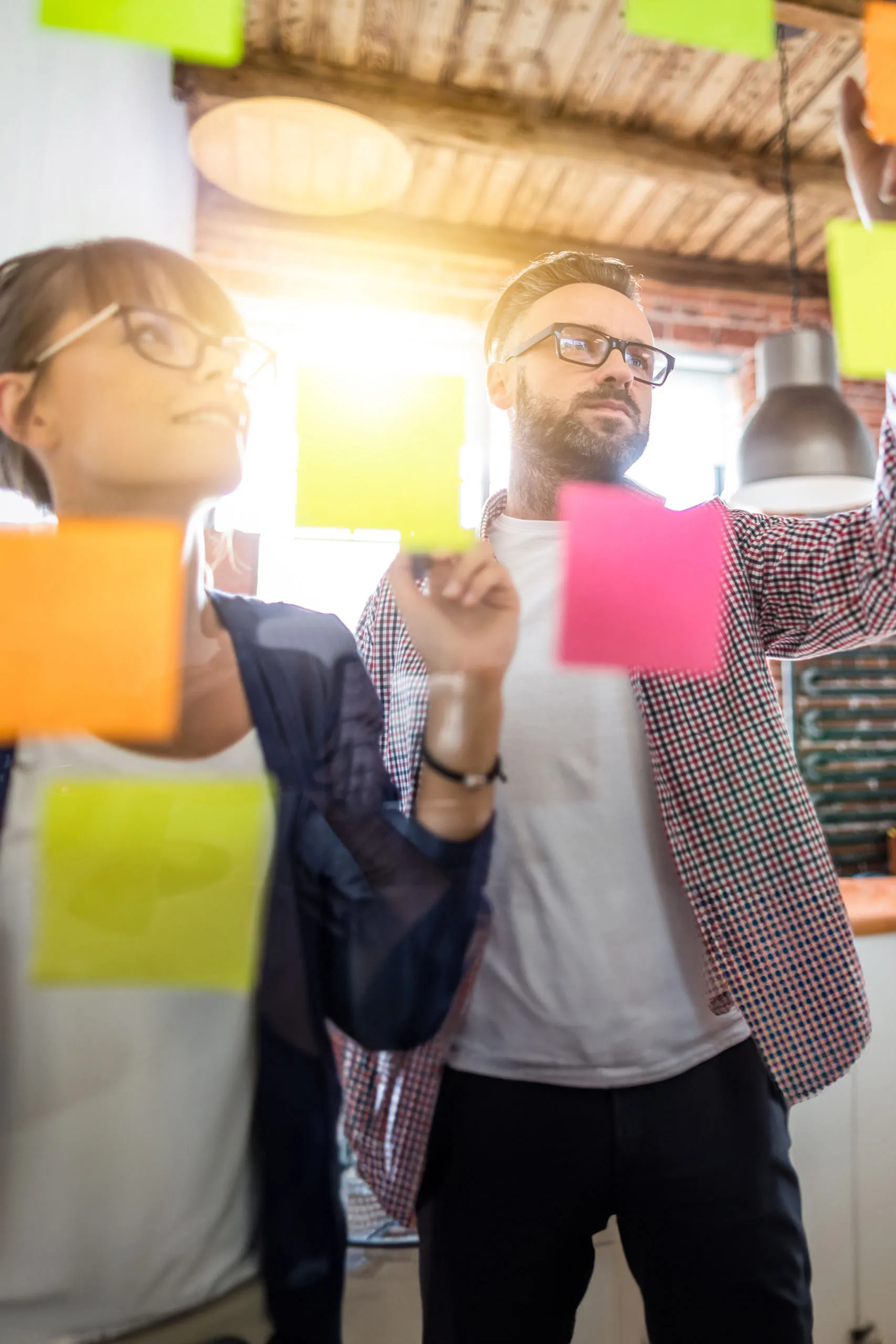 Two individuals collaborate, placing colorful sticky notes on a glass surface in a modern workspace.